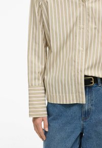 Chemise à manches longues beige à rayures vert clair et marron. Dotée de boutons sur le devant, d'un col et de larges poignets, portée avec un jean bleu.