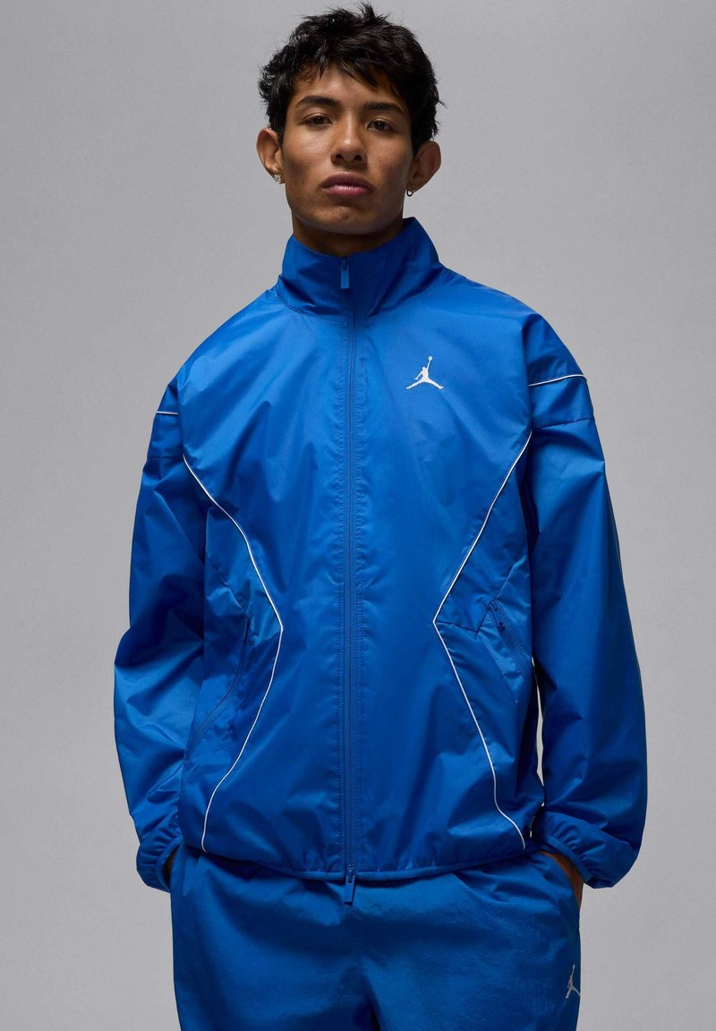 Blå vindjacka med vita detaljer, utrustad med hög krage, dragkedja framtill och elastiska muddar. Logotyp på övre bröstet, slät textur.