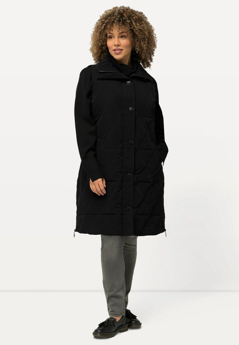 Mustat tikkivuorelliset takki napitussuljenta, vetoketjukaulus ja pitkät hihat, yhdistettynä harmaisiin housuihin ja mustiin kenkiin. Sujuva, teksturoitu kangas.