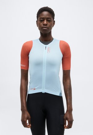 Μεγάλο άτομο που φοράει εφαρμοστή ποδηλατική φανέλα με ανοιχτό μπλε κορμό και πορτοκαλί κοντά μανίκια, στέκεται όρθιο απέναντι σε ένα απλό φόντο.