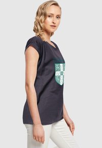 Mørkegrå t-shirt med rund halsudskæring, opsmøgede ærmer og et Cambridge University-våbenskjold i teal og hvid på forsiden.