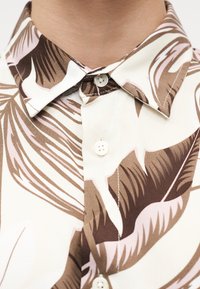 Γιακάς πουκάμισου μπεζ με κουμπιά, με καφέ και ανοιχτό ροζ τροπικό φυλλώδες μοτίβο, που δείχνει το πάνω μέρος του στήθους και την περιοχή του λαιμού.
