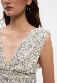 Srebrna haljina s šljokicama, dubokog V-izreza i nabranim rukavima. Glatka tekstura tkanine, prekrivena okruglim reflektirajućim šljokicama.