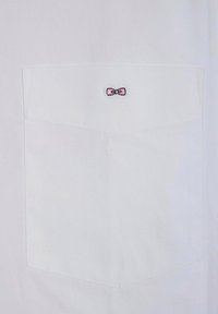 Chemise blanche avec une poche plaquée ornée d'une petite broderie en forme de nœud rose. Le tissu semble lisse avec un design épuré et ajusté.