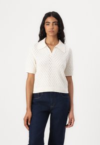 Short-sleeve, cream-colored knitted pullover with a diamond pattern and a wide collar, paired with dark blue denim pants.