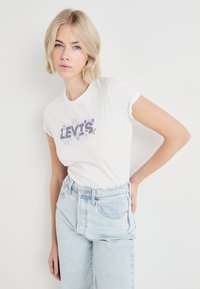 Biela bavlnená tričko s okrúhlym výstrihom, s fialovým logom "LEVI'S" obklopeným kvetinovými motívmi. V kombinácii so svetlom umytými nohavicami s vysokým pásom.
