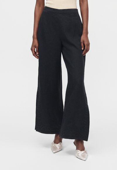 Personne portant un pantalon noir à jambes larges et des chaussures légères à bout pointu, sans lacets, debout devant un fond uni.