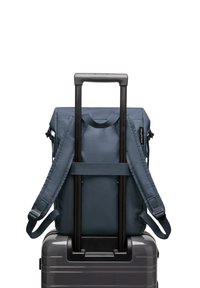 Sac à dos en tissu bleu marine avec des bretelles ajustables, posé sur une valise rigide argentée avec une poignée rétractable.