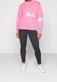 Sweat-shirt rose avec logo blanc "ELLESSE", associé à un legging noir et des baskets blanches. La tenue présente un style décontracté et sportif.