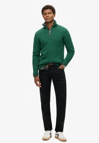 Grüner Strickjacke mit Reißverschluss und gerippten Bündchen, getragen über einem karierten Hemd, kombiniert mit schwarzen Jeans und weißen Sneakers mit schwarzen Akzenten.