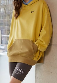 Gul hoodie med svart logotyp, tillsammans med bruna shorts som har vit text. Mjuk tyg, avslappnad passform och stor framficka.