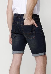 Pantalones cortos de mezclilla azul oscuro con puños enrollados, costuras en contraste y una pequeña etiqueta de tres colores en el bolsillo trasero. Dos bolsillos traseros visibles.