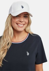 Căciulă albă cu un logo navy, purtată cu o cămașă navy care are un guler cu dungi roșii și albastre. Cămașa are un mic logo brodat în partea stângă.