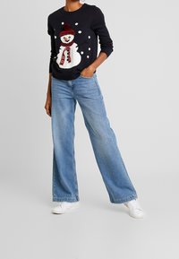 Persona che indossa un maglione nero con un design di un pupazzo di neve sorridente, jeans a gamba larga blu chiaro e sneakers bianche, in piedi davanti a uno sfondo uniforme.