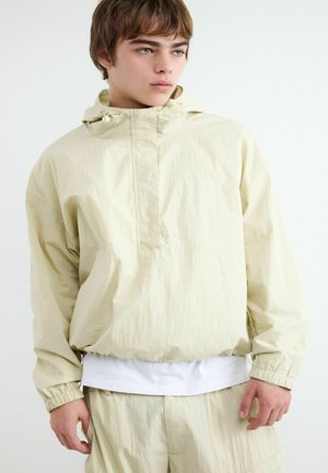 Młoda osoba w beżowej kurtce przeciwwiatrowej z kapturem, elastycznymi mankietami i pasujących spodniach, patrząca na bok na jednolitym białym tle.