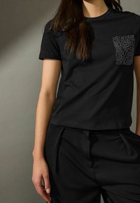 Sort bomulds t-shirt med korte ærmer, der har en tekstureret lomme med abstrakte mønstre. Sammen med sorte skræddersyede bukser.