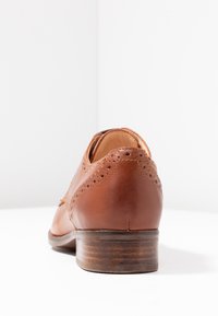Chaussure en cuir marron avec un design brogue, dotée d'un talon en bois et d'une texture lisse. La vue arrière met en avant des coutures détaillées et des perforations.