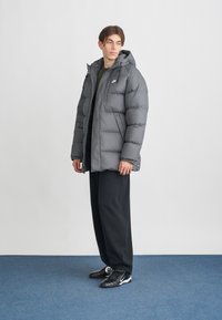 Casaco puffer cinza com capuz, textura acolchoada, fecho frontal e bolsos laterais. Usado sobre uma blusa escura e combinado com calças e sapatos pretos.