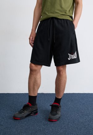 Man standing on blue carpet, wearing a green shirt and black basketball shorts with "JORDAN" printed, paired with black and red sneakers.