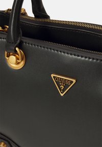 Sac à main en synthétique noir avec quincaillerie dorée, un logo en forme de triangle avec "GUESS USA" et une fermeture à glissière. Texture lisse avec un minimum de détails.