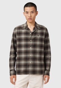 Man wearing brown and gray plaid button-up shirt with small embroidered logo, paired with light-colored pants, standing against plain background.