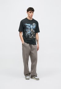 Schwarzes T-Shirt mit Grafikdesign, das Wölfe und Blitz zeigt, kombiniert mit grauer Jogginghose und grauen und weißen Turnschuhen.