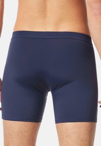 Man i tajta marinblå badshorts, visande bakifrån från midjan till mitten av låret mot en enkel vit bakgrund.