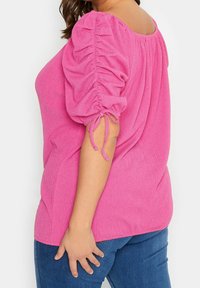 Růžový texturovaný top s vypuštěnými rukávy a šňůrkovými detaily na manžetách, v kombinaci s tmavě modrými džíny. Kulatý výstřih a volný střih.