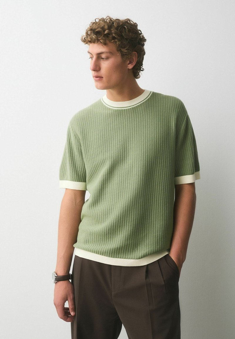 Jeune homme aux cheveux bouclés portant un pull en maille vert clair à manches courtes avec bordure blanche et un pantalon marron, debout contre un mur blanc uni.