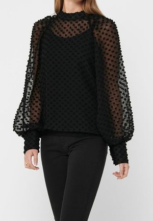 Mujer con blusa negra transparente de lunares, con mangas abullonadas y cuello alto, combinada con pantalones negros ajustados.