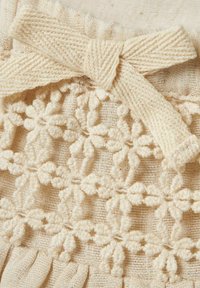 Beige texturerad tyg med en vävd blommig spetsdetalj och en rosett högst upp. Innehåller delikat, intrikat sömnad genom hela tyget.