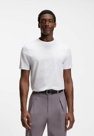 Uomo in piedi su sfondo bianco che indossa una semplice maglietta bianca a maniche corte infilata in pantaloni grigi plissettati con cintura nera.