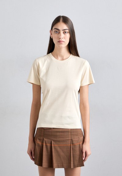 Frau mit langen braunen Haaren, die ein cremefarbenes T-Shirt mit kurzen Ärmeln und einen braunen plissierten Rock mit orange und schwarzem Karomuster trägt.
