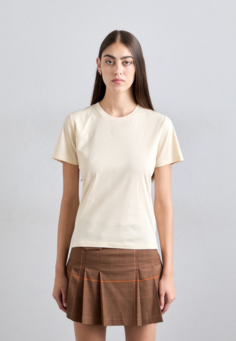 Woman with long brown hair wearing a cream short-sleeve t-shirt and a brown pleated skirt with orange and black plaid pattern.