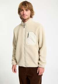 Jeune homme aux cheveux blonds ébouriffés portant une veste polaire beige avec une poche poitrine zippée et un pantalon marron sur fond blanc uni.