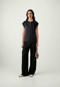 Black short-sleeved top with a front keyhole detail, paired with high-waisted black trousers and white slip-on shoes, carrying a small brown bag.
