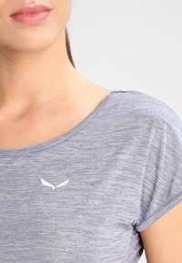 Světle šedý top s krátkým rukávem vyrobený z měkké, texturované látky. Má malé bílé logo na hrudi a kulatý výstřih.