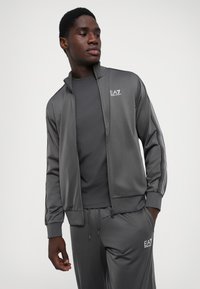 Chaqueta deportiva gris con cuello alto y cremallera, combinada con una camiseta gris a juego. Presenta un logo de marca y acentos a rayas en las mangas.