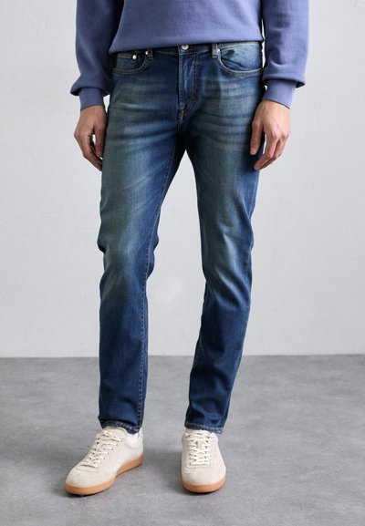 Jeans din denim albastru pentru bărbați, cu o croială slim, spălare ştearsă, design cu cinci buzunare și accesorii metalice, asortați cu adidași în culori deschise.