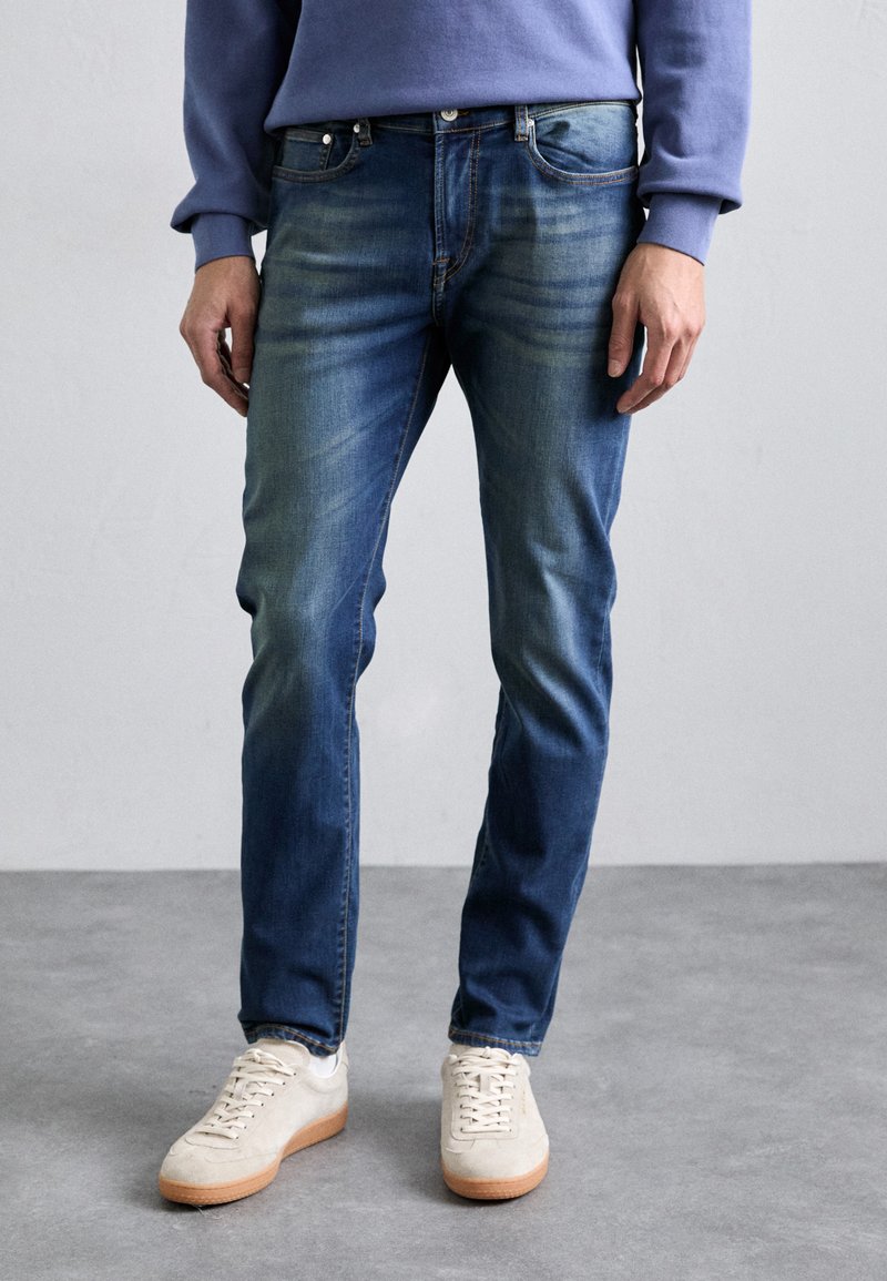 Férfi kék farmernadrág slim fit szabással, koptatott mosással, öt zsebbel és fémből készült kiegészítőkkel, világos színű sneakers cipőkkel párosítva.