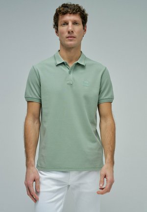 Hombre vistiendo una camisa polo de manga corta lisa de color verde claro y pantalones blancos, posando frente a un fondo gris liso.