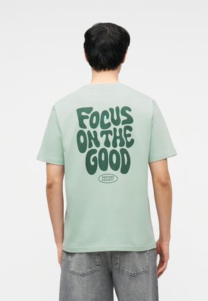 Személy világos zöld pólót visel sötétzöld felirattal: "FOCUS ON THE GOOD" és apró "KAOTIKO SOCIETY" logóval a hátoldalon.