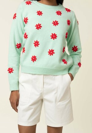 Vaaleanvihreä neule, jossa punaiset kukkakoristeet, yhdistetty valkoisiin polvenmittaisiin shortseihin, mallin oikea käsi taskussa, neutraali tausta.