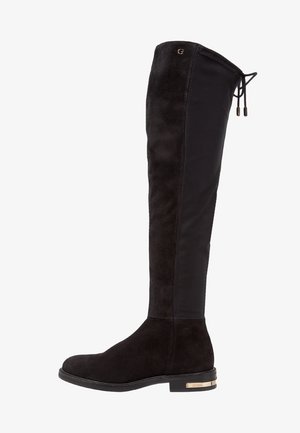 Bottes noires au-dessus du genou avec un devant en daim et un dos élastique, ornées d'un logo doré sur le talon et d'une semelle plate.