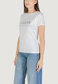 T-shirt bianca in cotone con maniche corte, con il logo "GUESS" in rilievo argentato sulla parte anteriore. Design aderente con una texture liscia.
