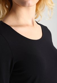 Svart, åtsittande topp med rundad halsringning, tillverkad av slät tyg. Närbild som framhäver tygtexturen och halsringningens design.