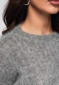 Partie inférieure du visage et cou d'une femme portant une boucle d'oreille en diamant et un pull gris en maille douce avec un motif torsadé.