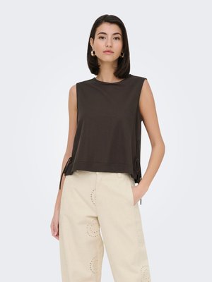 Femme aux cheveux courts et foncés porte un haut noir sans manches et un pantalon beige clair avec des détails de perforations circulaires, une main dans la poche.