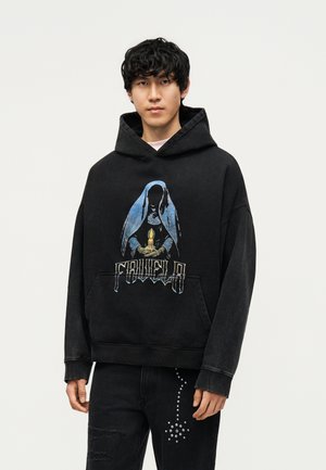 Jonge man met zwart krullend haar die een zwarte hoodie draagt met een blauw-gekapte figuur die een brandende kaars vasthoudt en zwarte jeans met een met studs versierd kruisontwerp.