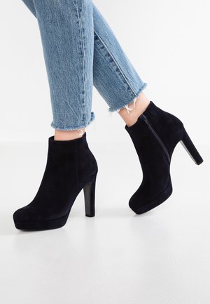 Personne portant un jean bleu à ourlet effiloché et des bottines noires en daim à talons hauts avec fermetures éclair latérales sur une surface blanche.